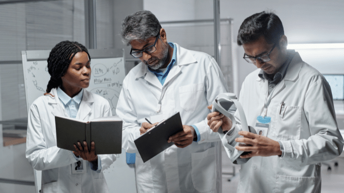 Three scientists in white lab coats stand together in a lab discussing their work. One woman holds an open notebook, one man writes on a clipboard, and another man examines a grey VR headset.