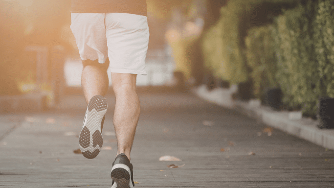 A shot from behind of a person jogging down a paved path lined with green hedges. The scene is bathed in warm, soft sunlight, emphasizing a healthy, active morning routine.