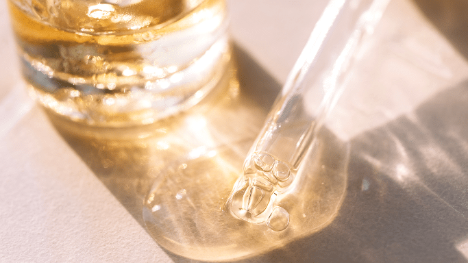 A close-up, warm-toned shot of a glass pipette dispensing a thick, clear cosmetic oil or serum onto a surface next to a glass bottle. The scene is illuminated by soft, golden sunlight.