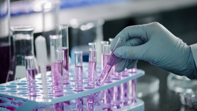 A laboratory setting where a gloved hand is carefully selecting a glass test tube filled with purple liquid from a blue rack. Other lab glassware, including beakers and flasks, is blurred in the background.