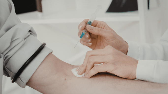 A close-up of a medical professional’s hands wearing white sleeves, preparing to draw blood or administer an injection into a patient's arm using a syringe and cotton swab.