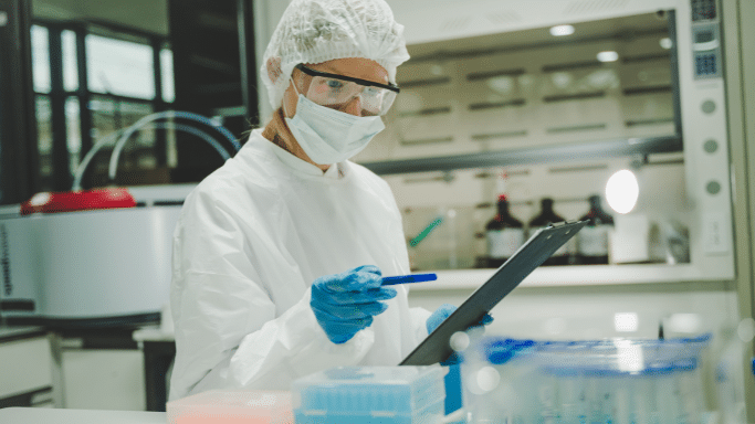 A scientist wearing a full white protective suit, hairnet, face mask, and blue gloves looks intently at a clipboard while holding a blue pen. The background shows a sterile laboratory with a fume hood and various containers.