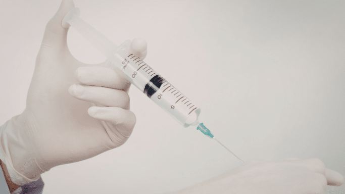 A close-up of a hand in a white latex glove holding a large medical syringe filled with clear liquid, positioned as if ready for an injection against a plain, light grey background.