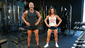 A fit middle-aged man and woman stand side-by-side in a well-equipped gym. The man wears a black tank top and shorts, showcasing muscular legs, while the woman wears a white tank top and blue shorts. Both are smiling with their hands on their hips.