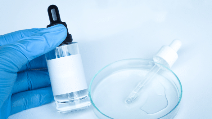 A close-up of a hand in a blue nitrile glove holding a small glass dropper bottle with a blank white label. In the background, a glass petri dish sits on a white surface with a dropper and a small pool of clear liquid.