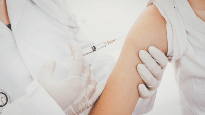 A close-up, high-angle shot of a medical professional in a white coat and gloves administering an injection into a patient’s upper arm.