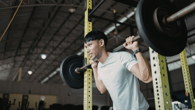 A man in a white t-shirt performs a barbell back squat inside a yellow power rack in a gym.