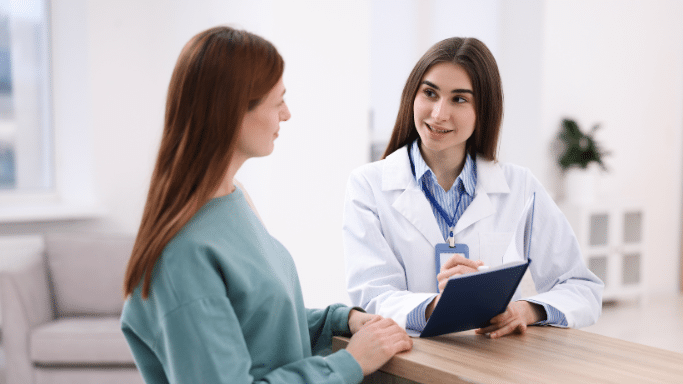 A female healthcare professional in a white lab coat and blue striped shirt talks to a female patient. She is holding a blue clipboard and a pen, appearing to explain or record medical information in a bright clinic setting.