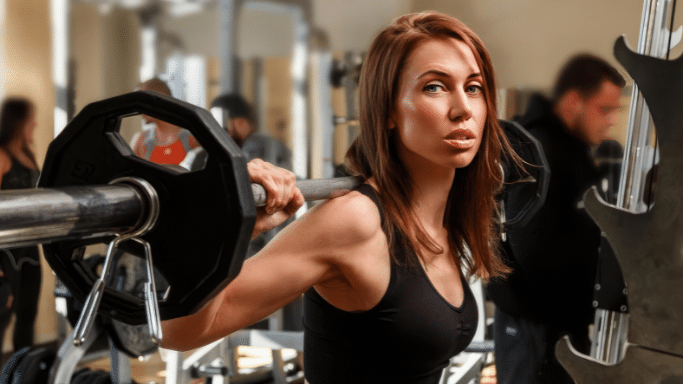 A woman with reddish hair looks forward while holding a weighted barbell across her shoulders for squats.