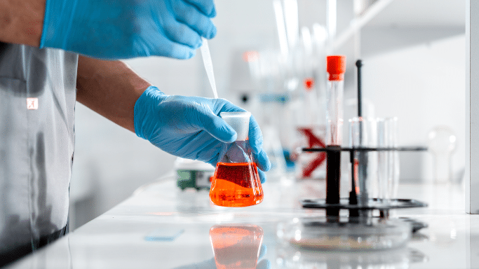 Close-up of gloved hands using a pipette to add liquid to an Erlenmeyer flask containing orange fluid.