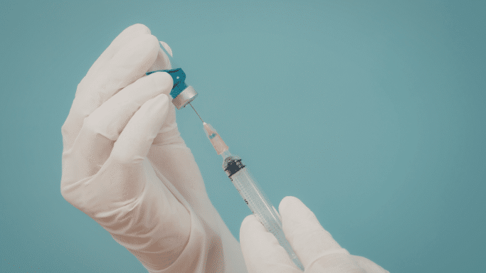 A close-up of gloved hands carefully drawing liquid from a small glass vial into a syringe against a plain teal background.