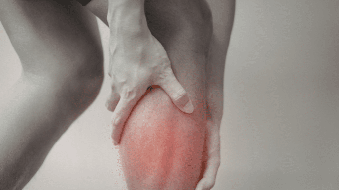 A black-and-white photo with a selective red highlight on a person's calf muscle. The person's hands are gripping the leg, indicating a point of pain, strain, or injury.