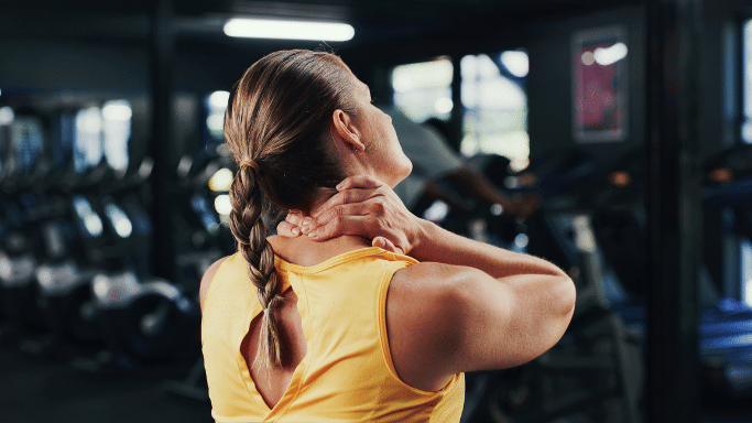 Rear view of a woman in a gym holding her neck, appearing to be in pain or stretching.