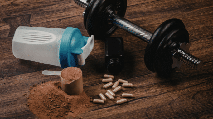 Fitness supplements, including protein powder, capsules, a shaker bottle, and a dumbbell, on a wooden surface.