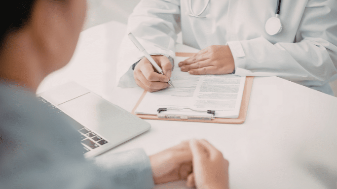 A first-person view looking across a white desk at a doctor. The doctor’s hands are visible as they write on a medical form attached to a clipboard. A laptop and the patient's clasped hands are also visible on the desk.
