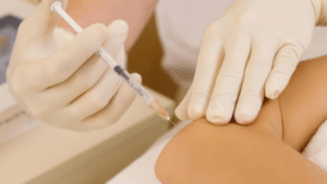 A close-up shot showing a pair of hands in white medical gloves. One hand holds a small syringe with a needle, while the other hand rests near a person's elbow, preparing the skin for an injection.