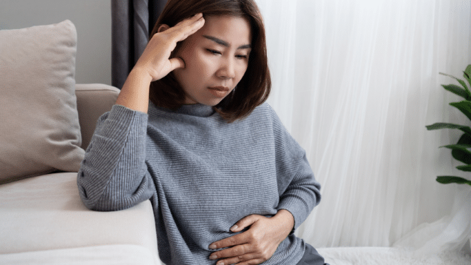 A woman sits on the floor leaning against a sofa, looking distressed. She has one hand on her forehead and the other on her abdomen, suggesting she is experiencing both a headache and stomach pain.