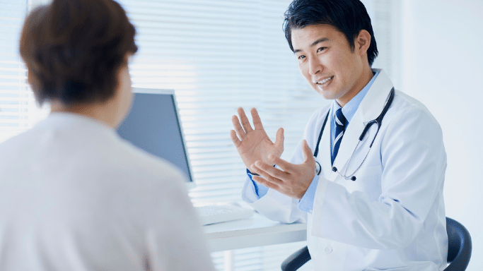 A male doctor in a white lab coat and stethoscope sits at a desk, gesturing with both hands while speaking to a patient, who is seen from the back. The setting is a bright, professional medical office.