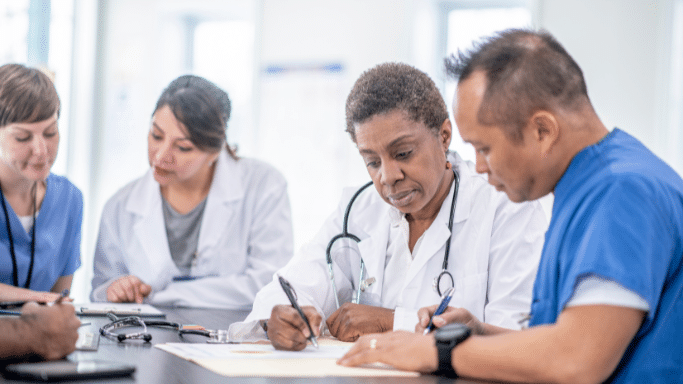 A diverse group of four medical professionals in lab coats and scrubs sit around a table in a meeting. They are focused on paperwork, with a stethoscope lying on the table in front of them.