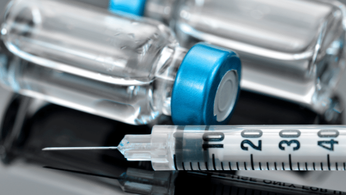 A macro shot showing a medical syringe with a sharp needle lying in front of two glass medicine vials with blue caps. The surface beneath them is reflective.