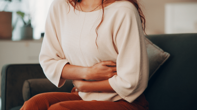 A woman in a cream-colored sweater sits on a dark green couch, clutching her stomach with both hands, indicating abdominal discomfort or cramping.