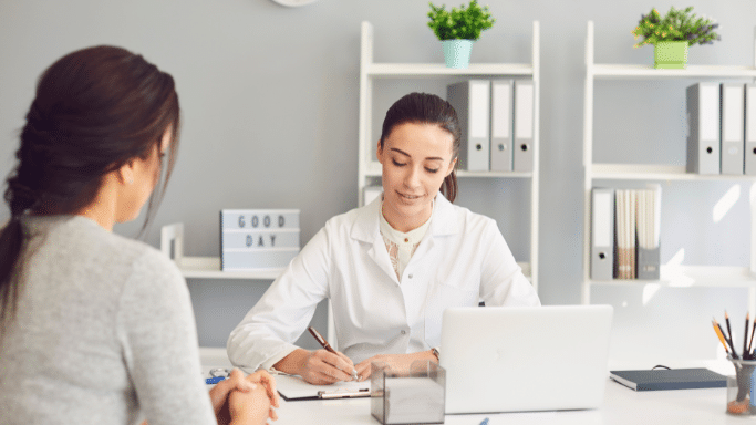 A female doctor sits at a white desk with a laptop, writing on a clipboard while consulting with a patient seated across from her. The background features clean, organized office shelving.