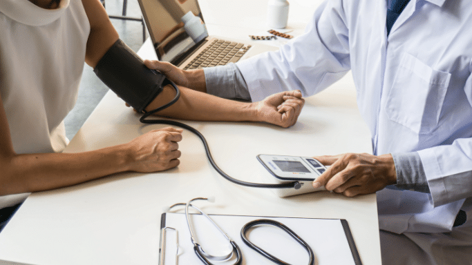 An over-the-shoulder view of a doctor performing a blood pressure check on a patient. The doctor’s hand is on the patient's arm near the cuff, while the other hand operates a digital blood pressure monitor on the desk.