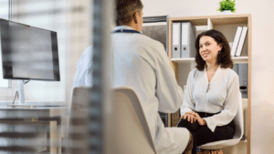 Endocrinologist speaking with a patient in a modern metabolic clinic Large screen shows a risk vs benefit framework (no dosing numbers): GI system risk markers (gallbladder/pancreatic icons in subtle warning tone) muscle mass preservation indicator dosing consistency timeline