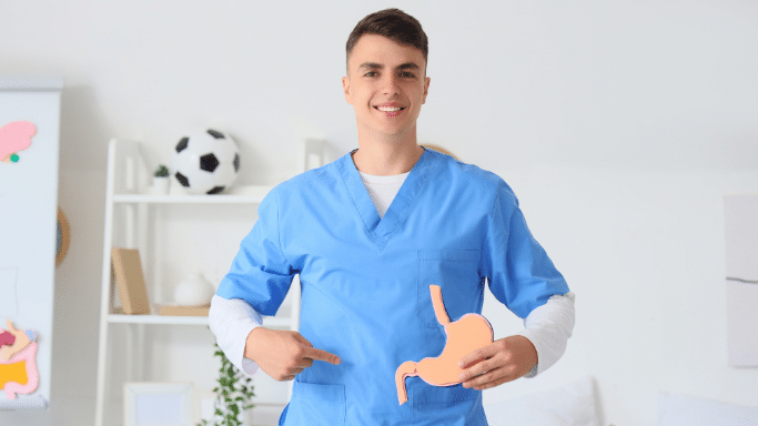 A young man in blue scrubs stands in a bright room, smiling and holding a flat, orange cutout model of a human stomach against his midsection. A soccer ball and shelves are visible in the background.