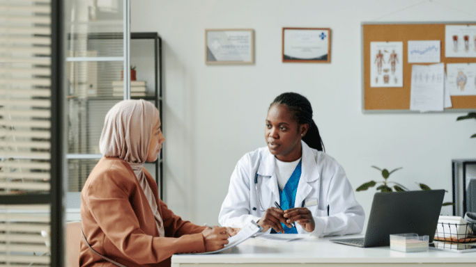 A Black female doctor in a white lab coat and blue scrubs sits at a desk, speaking with a patient wearing a tan hijab and blazer. The doctor holds a pen and looks focused on the conversation.
