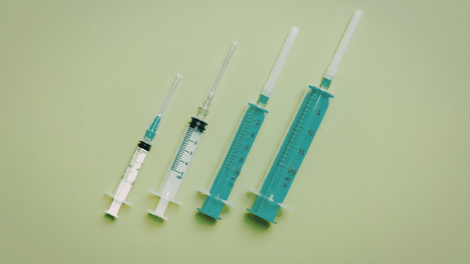 A top-down view of four medical syringes of increasing sizes laid out diagonally on a solid light-green background. Each syringe is capped and has clear measurement markings.