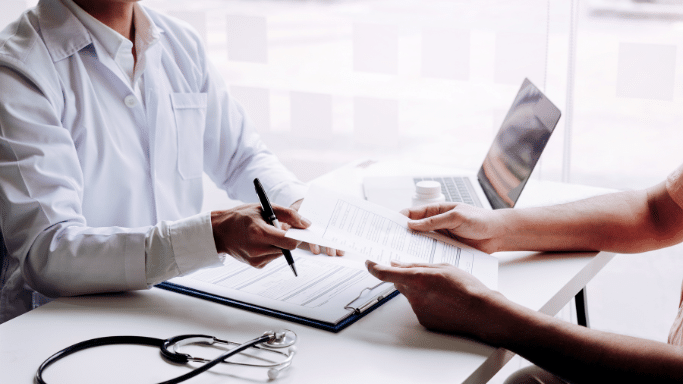 A close-up shot of a doctor's and patient's hands over a desk. The doctor is holding a pen and pointing at a document on a clipboard, while the patient holds the other side of the paper. A stethoscope and a laptop are visible on the white desk.