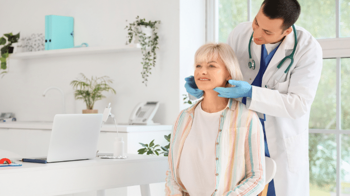 A male doctor wearing blue gloves stands behind a seated woman with blonde hair, gently performing a physical examination of her neck or thyroid area. They are in a bright, modern clinic.
