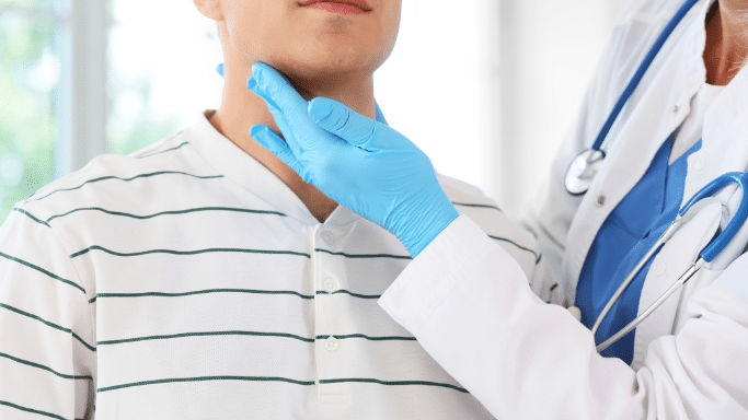 A close-up of a healthcare provider's hands in blue surgical gloves as they palpate the neck of a young man wearing a striped shirt.