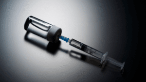 A close-up shot of a clear glass medical vial with a white cap lying horizontally on a dark, reflective surface. A plastic syringe with a blue needle hub is inserted straight into the rubber stopper of the vial, casting a prominent shadow underneath.