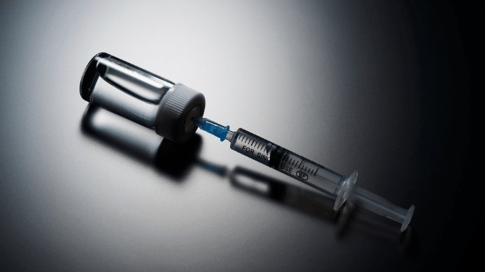 A close-up shot of a clear glass medical vial with a white cap lying horizontally on a dark, reflective surface. A plastic syringe with a blue needle hub is inserted straight into the rubber stopper of the vial, casting a prominent shadow underneath.