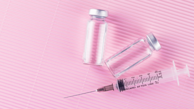 A top-down shot of two clear, empty medical vials with silver caps resting on a textured, light-pink background. Below the vials lies a clear plastic medical syringe with a thin needle, featuring black measurement markings and the text "DISCARD AFTER USE."