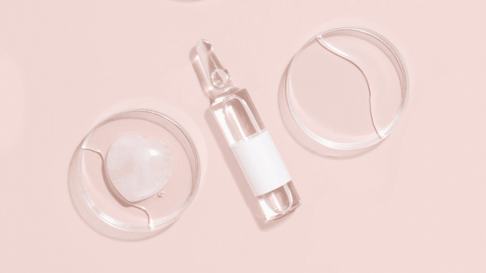 A minimalist, top-down product shot against a soft pink background. A clear glass medical ampoule with a blank white label is positioned diagonally in the center, flanked by two clear petri dishes—one on the left containing a light pink, heart-shaped stone submerged in liquid, and one on the right containing a simple fluid swirl.