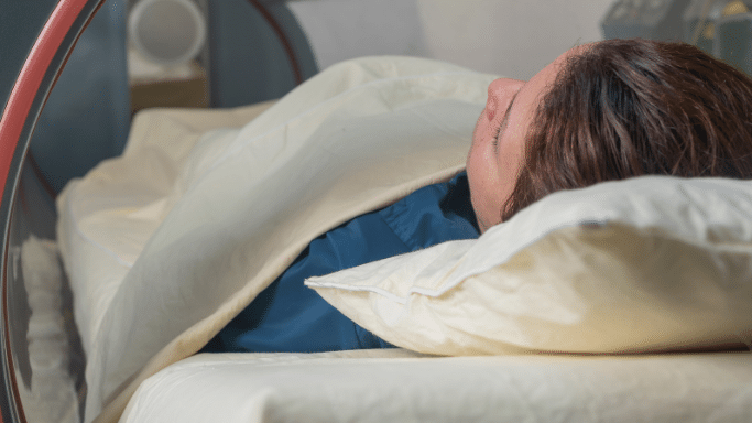 A close-up profile shot of a person lying down inside a hyperbaric oxygen chamber or medical capsule. The individual is covered up to their chest with an off-white blanket, resting their head on a matching pillow, viewed through the clear circular viewing window of the machine.