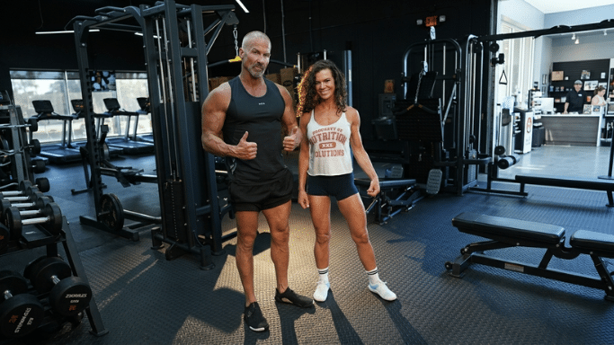 A full-body shot of a muscular man and woman standing together in a spacious, modern gym with dark grey walls and black rubber flooring. The man on the left wears a black tank top and black shorts, giving a thumbs-up. The woman on the right wears a white tank top that says "NUTRITION SOLUTIONS" and black shorts, flexing one arm. Rows of treadmills and strength equipment fill the background.