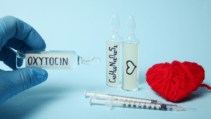 A close-up shot against a light blue background shows a gloved hand holding a glass ampoule labeled "OXYTOCIN." Behind it stand two other ampoules—one labeled with the chemical formula $\text{C}_{43}\text{H}_{66}\text{N}_{12}\text{O}_{12}\text{S}_{2}$ and another marked with a simple heart drawing. Two syringes lie flat in front of them next to a red yarn heart.