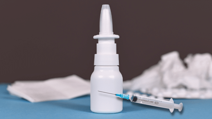 A white plastic nasal spray bottle stands upright on a blue surface against a dark gray background. A clear medical syringe with a blue needle hub rests diagonally in front of the bottle's base.