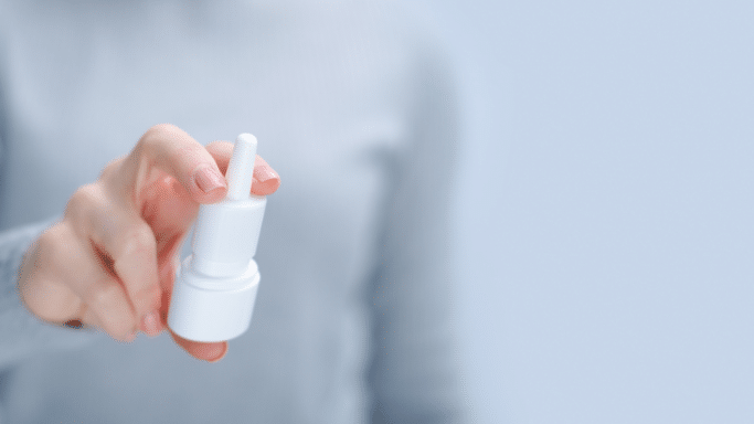 A close-up, slightly blurred shot of a person's hand holding a white nasal spray bottle, positioned as if ready to press down and dispense a dose.