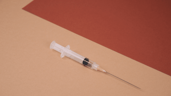 A high-angle shot of a clear plastic medical syringe with a long needle resting diagonally across a background divided into two solid colors: a light tan lower-left half and a dark reddish-brown upper-right half.