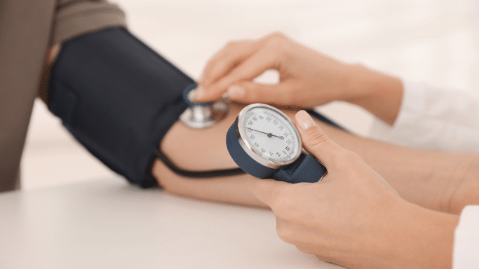 A close-up view showing a healthcare provider's hands checking a patient's blood pressure using a manual aneroid sphygmomanometer gauge and an arm cuff.