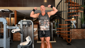 A muscular older man in a camo "IBIZA 87" tank top and shorts stands in a gym, flexing both of his biceps in a double-bicep pose. A wooden spiral staircase is visible on the right.