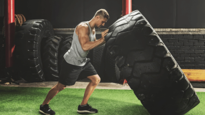 A side view of a muscular man in a gray tank top and black shorts flipping a massive, heavy tractor tire on a green turf floor inside a gym.