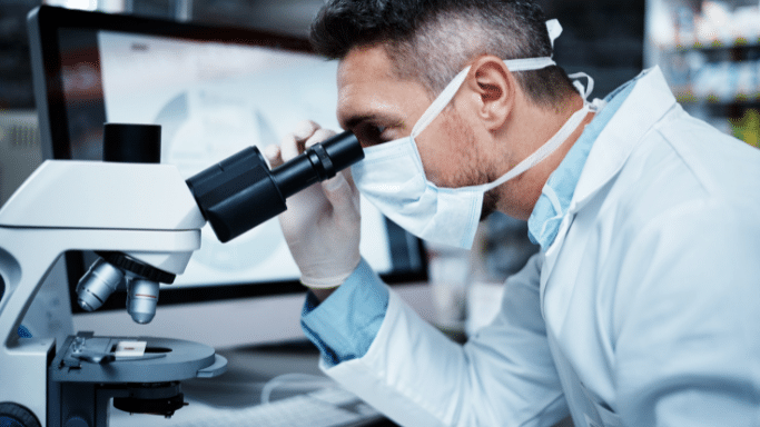 A side-profile shot of a scientist wearing a laboratory coat, hairnet, and a light blue surgical mask, intently looking into the eyepiece of a modern microscope. A computer monitor showing a blurry chart is visible in the background.