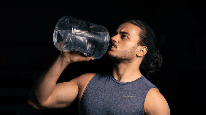 A man with dark hair tied back in a bun, wearing a gray sleeveless Nike shirt, drinking water from a large, clear gallon jug against a dark, minimalist background.