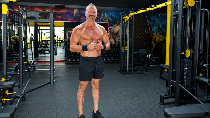 A smiling, muscular older man with a mohawk hairstyle and wrist wraps stands in the middle of a spacious, well-equipped gym with black and yellow workout machines. He is shirtless, wearing black shorts, and giving a thumbs-up.